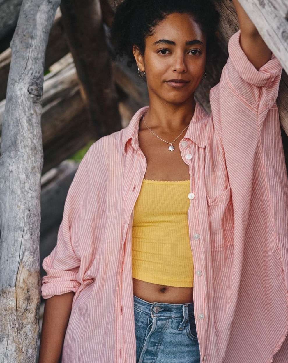 A young woman with curly dark hair poses outdoors wearing a pink button-down shirt, yellow top, and denim shorts, smiling at the camera.