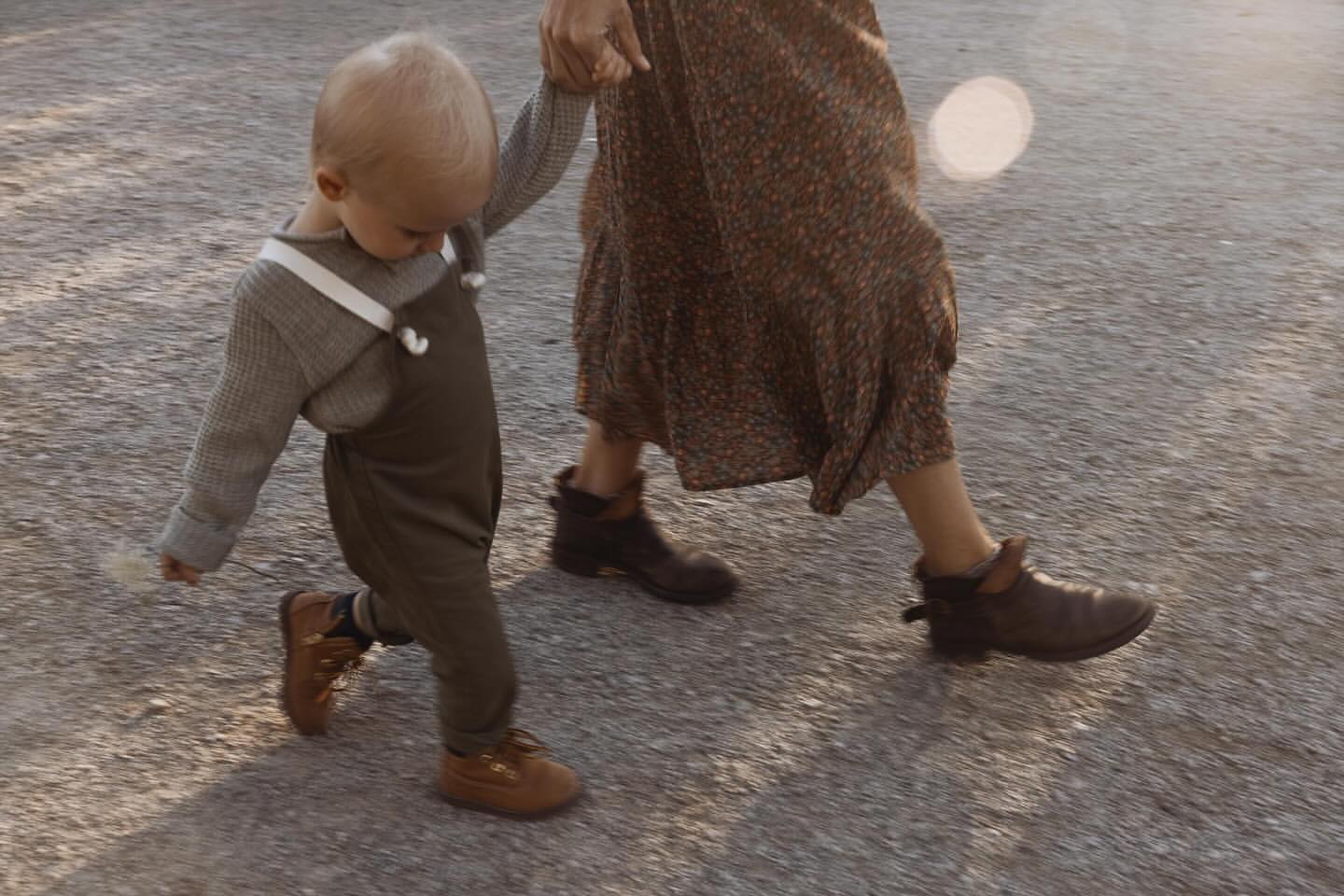 Low-angle shot of a toddler holding an adult's hand walking outside on a sunny day. The child wears overalls and brown shoes.