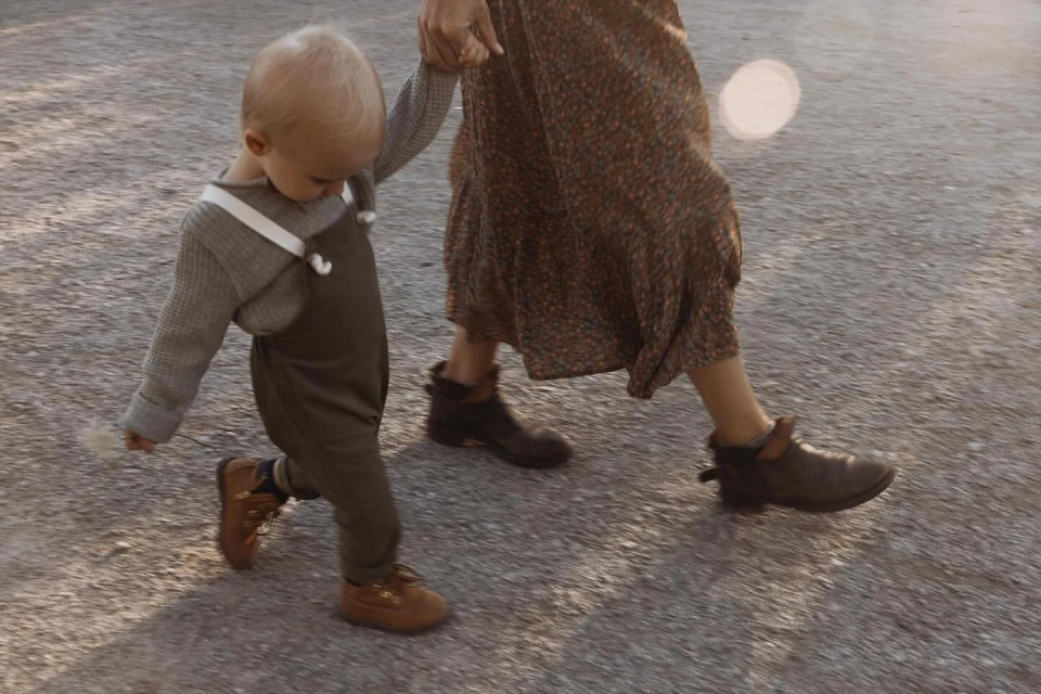 Low-angle shot of a toddler holding an adult's hand walking outside on a sunny day. The child wears overalls and brown shoes.