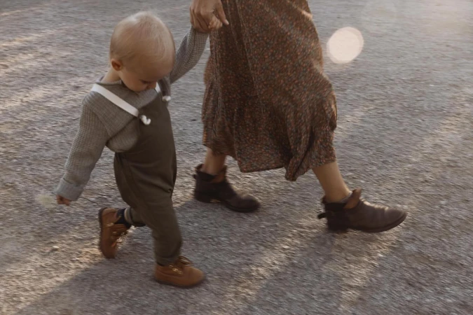 Low-angle shot of a toddler holding an adult's hand walking outside on a sunny day. The child wears overalls and brown shoes.