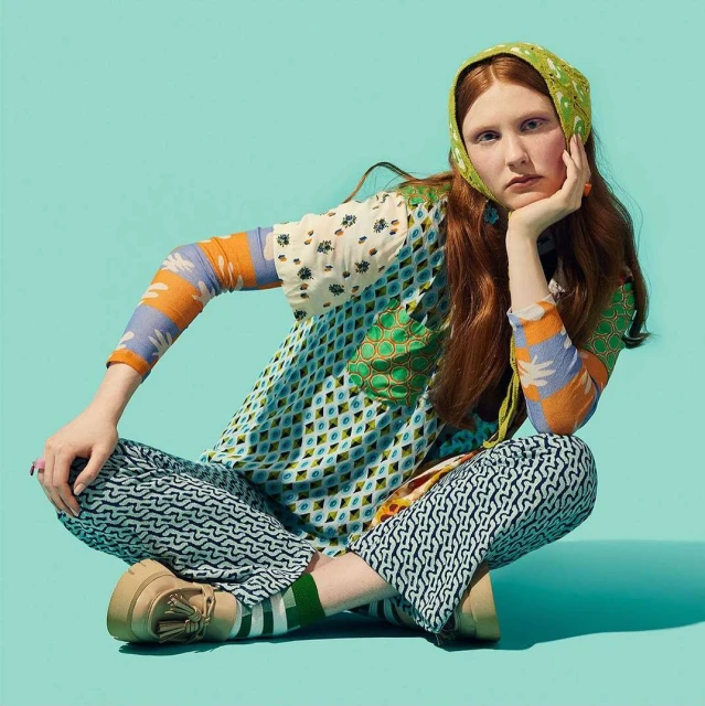 Red-haired woman in patterned clothing sits cross-legged, head resting on hand against a vibrant teal backdrop in a fashion editorial.