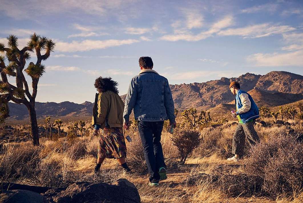 Three people walking away on a desert trail with Joshua trees. Mountains are in the background under a blue sky with clouds.