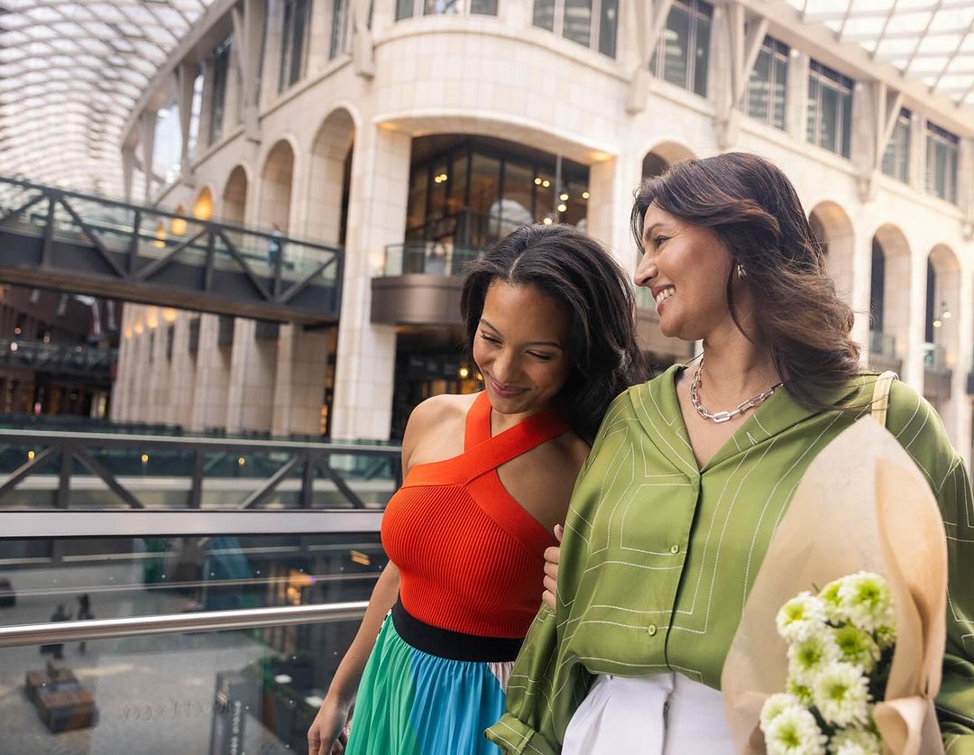 Two women walk arm-in-arm in a sunny shopping mall. One carries flowers. A bridge and beige building visible in the background.