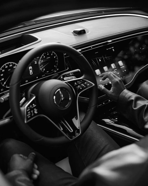 Interior view of a Mercedes-Benz dashboard featuring a steering wheel, digital displays, and a person's hand interacting with the touchscreen.
