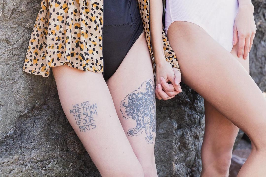 Close-up of two women holding hands, featuring leg tattoos with one woman in a cheetah-print top leaning against a rock wall.