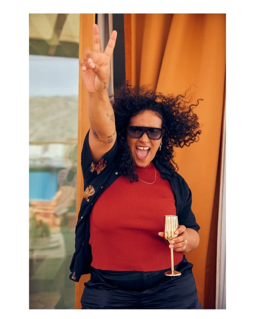 A cheerful woman with curly hair raises her hand in a peace sign gesture while holding a champagne flute. She is wearing sunglasses.