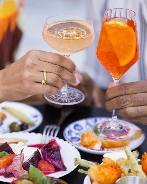 Two people toasting cocktails. Clear glasses hold colorful drinks garnished with citrus slices, plates of food on dark table.