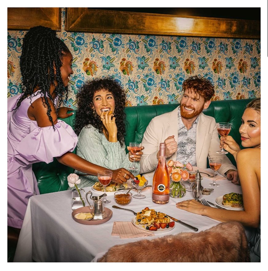 Five smiling people at a restaurant table with food, drinks, and floral wallpaper in the background during daytime.
