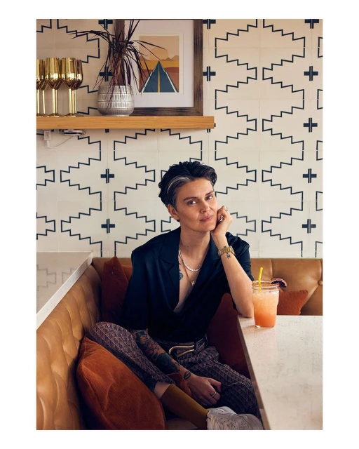 Stylish woman in a black blazer sitting indoors on a leather banquette with a colorful drink, in front of a geometric pattern wall.