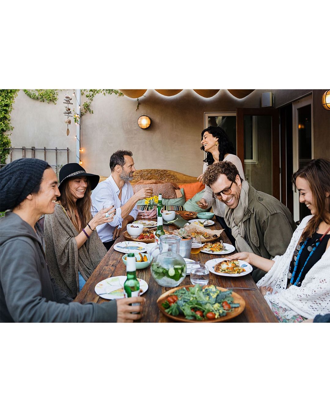 Group of six people seated at a wooden table, sharing food and drinks outdoors in a well-lit patio setting, with greenery in the background.