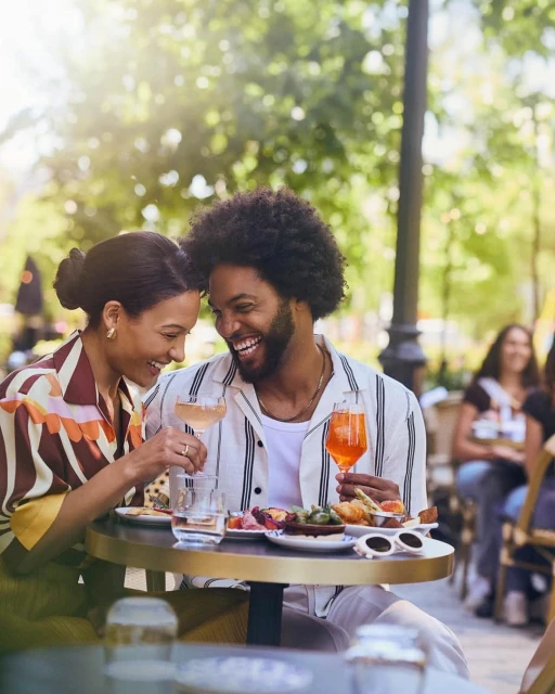 A happy couple enjoys food and drinks at an outdoor café on a sunny day, with the background blurred to emphasize the foreground.