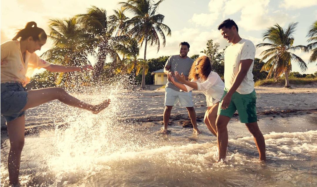 A group of four people, including two women and two men, are playing in the ocean surf under a sunny sky near a sandy beach.