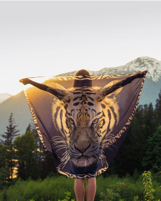 Person stands with outstretched arms while holding a tiger print shawl, with a view of distant, green mountains in the background.
