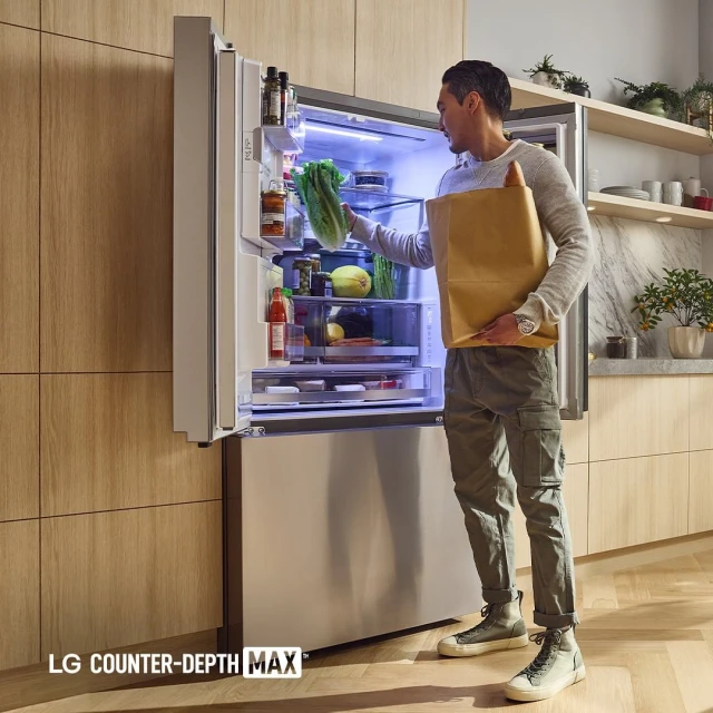 Adult male places fresh lettuce into a stainless steel LG refrigerator filled with various food items in a light and airy kitchen.