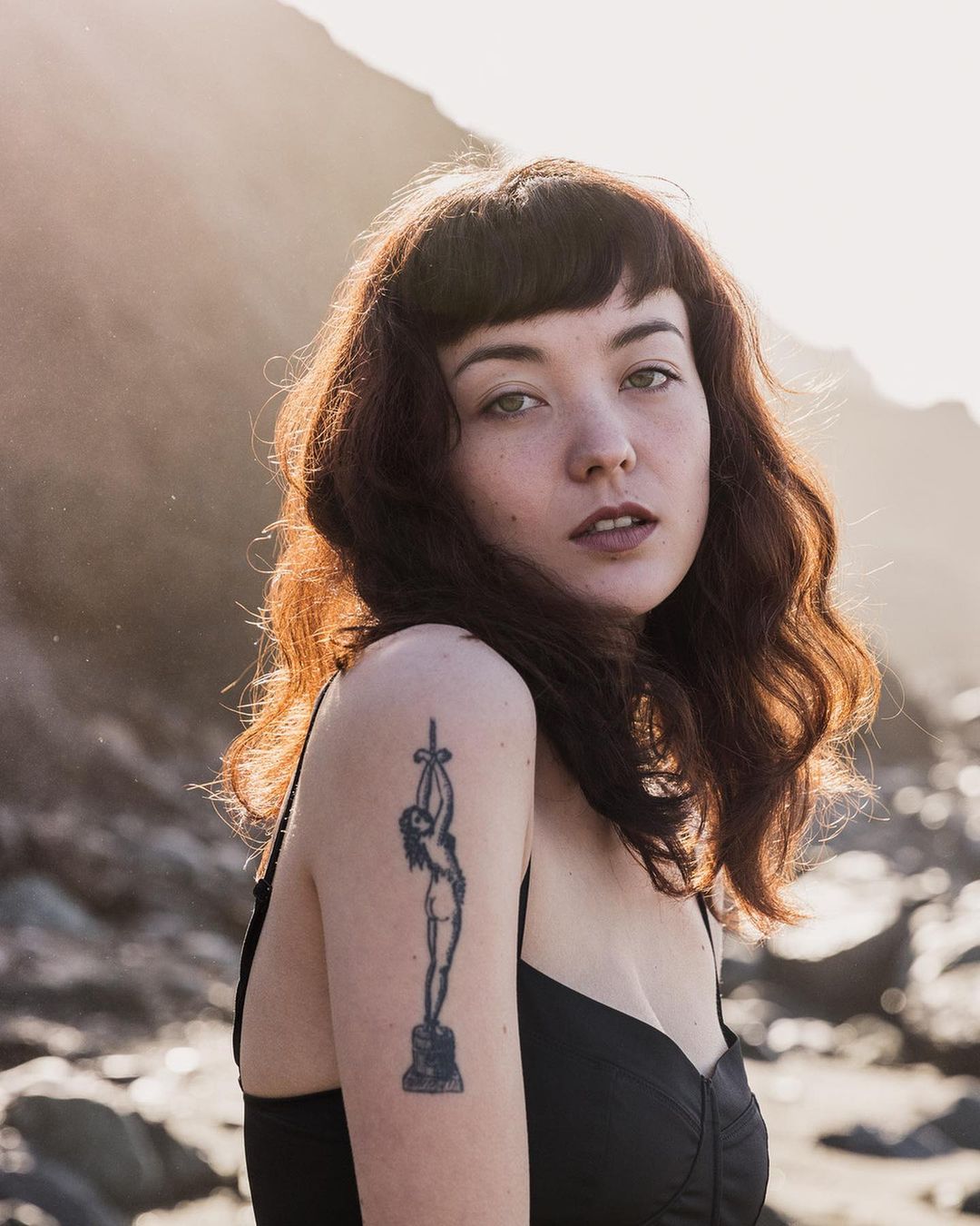 Portrait of a woman with dark wavy hair and bangs wearing a black top, showing a detailed tattoo on her shoulder outdoors.