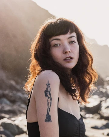 Portrait of a woman with dark wavy hair and bangs wearing a black top, showing a detailed tattoo on her shoulder outdoors.