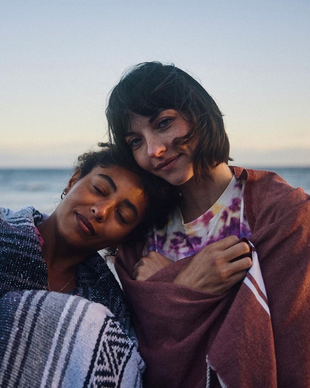 Two women embrace on a beach at sunset, wrapped in colorful blankets, showcasing a peaceful and intimate moment by the ocean.