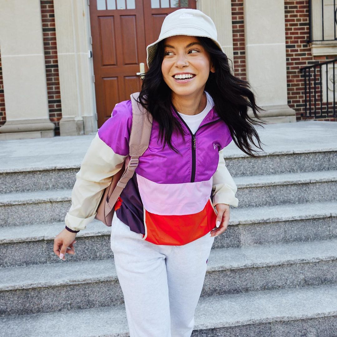 Smiling woman in a colorful jacket and bucket hat stands on stone steps with a backpack in front of a brick building in daylight.