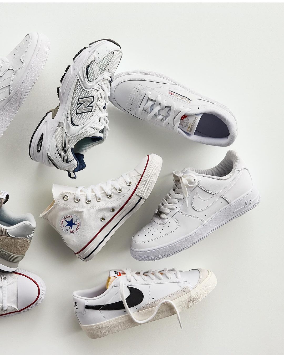 Collection of athletic shoes in white and neutral tones including sneakers and canvas trainers, artfully arranged on a light background.