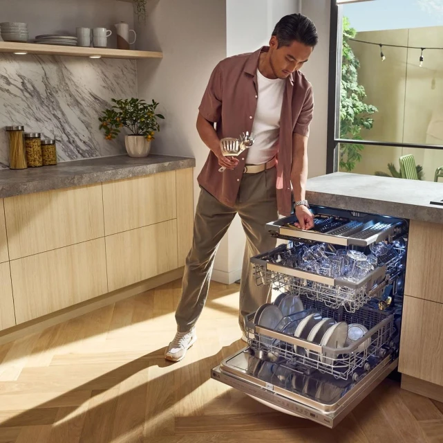 Adult male stands in a modern kitchen, loading silverware into the top rack of an open, stainless steel dishwasher with interior lighting.