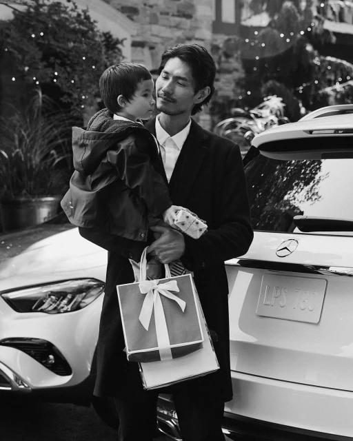 Black and white portrait of Asian father holding his young child near a white Mercedes SUV, standing outdoors near a stone building.