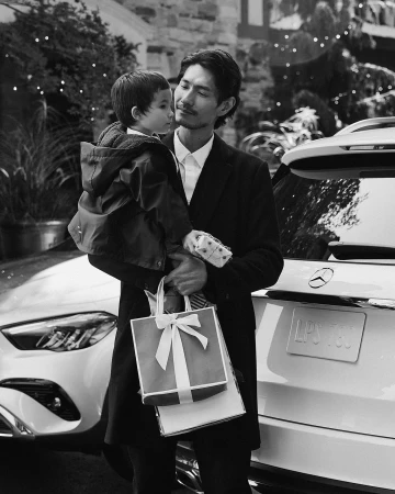 Black and white portrait of Asian father holding his young child near a white Mercedes SUV, standing outdoors near a stone building.
