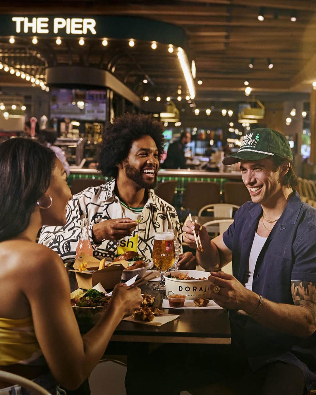 Three friends laugh while eating takeout food together at a restaurant with overhead lighting. The food includes fries and fried chicken.