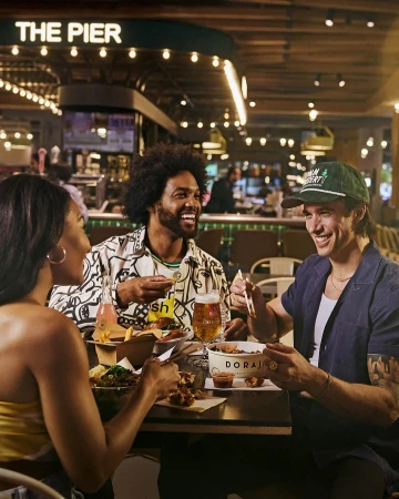 Three friends laugh while eating takeout food together at a restaurant with overhead lighting. The food includes fries and fried chicken.