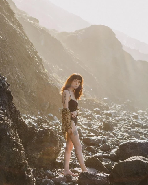 Woman in a swimsuit and leopard print cardigan stands on a rocky beach with a mountain backdrop during golden hour. Portrait orientation.