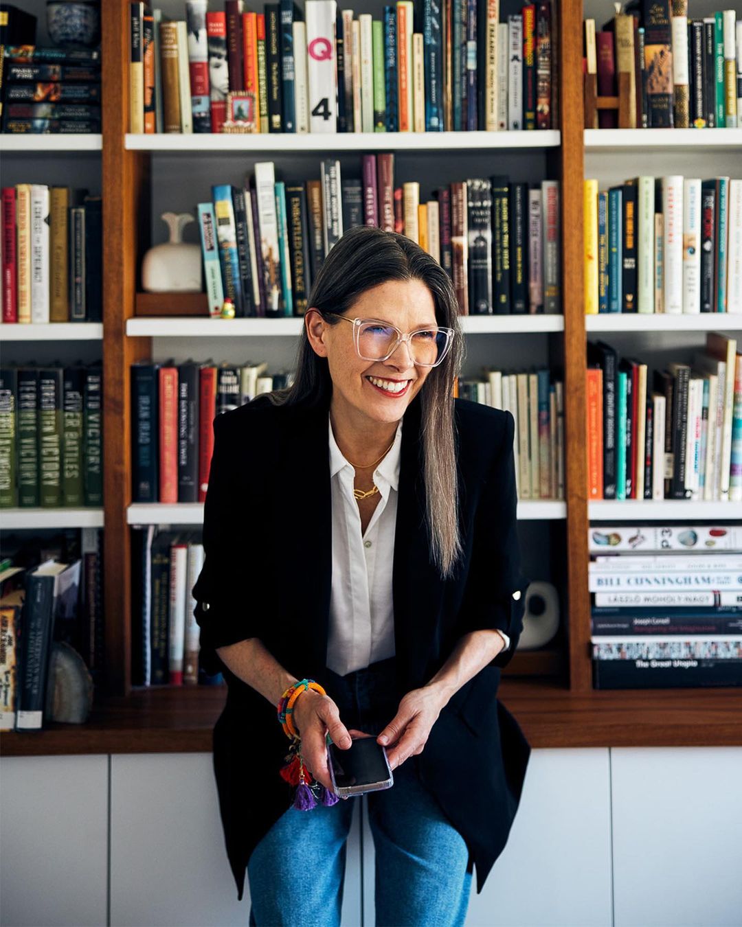 Adult woman smiles and poses indoors near a bookshelf in front of a white wall, wearing a blazer, glasses, and holding a phone.