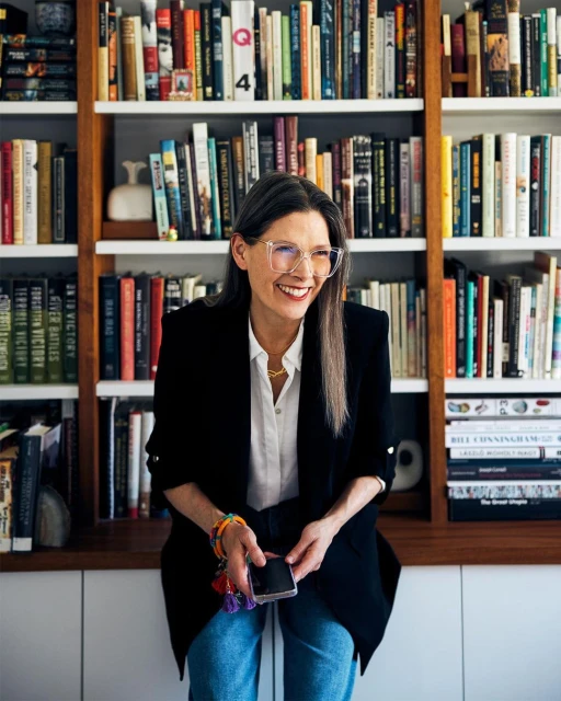 Adult woman smiles and poses indoors near a bookshelf in front of a white wall, wearing a blazer, glasses, and holding a phone.