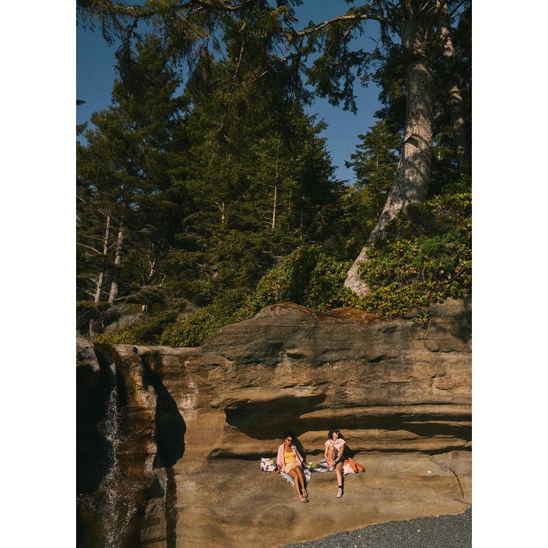 Two women relax on rocks at a beach near a forest on a sunny day. Tall trees frame the scene, providing a lush, natural backdrop.