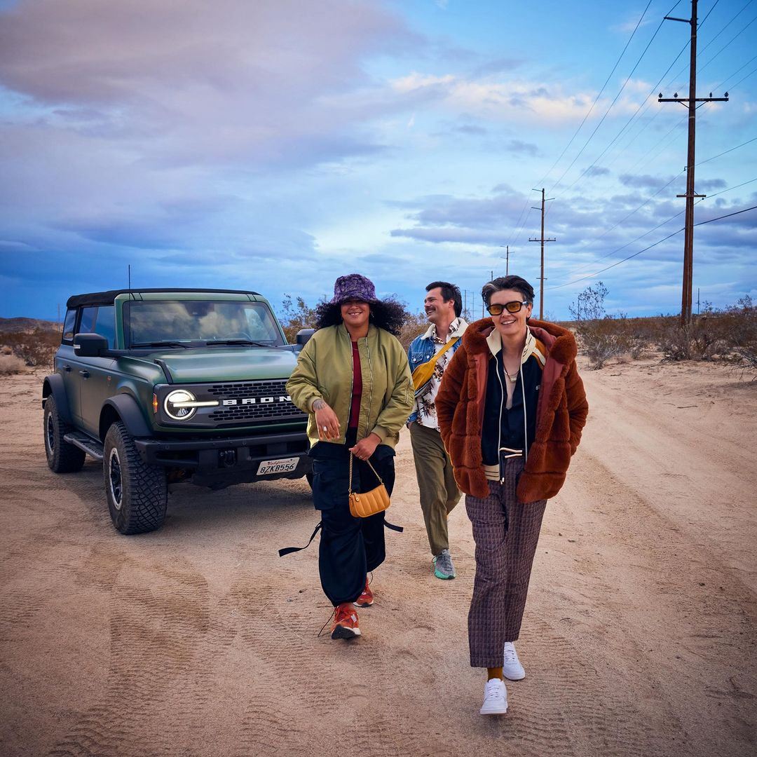 Three people walking toward the camera in the desert with a Ford Bronco SUV in the background, under a cloudy sky at dusk.