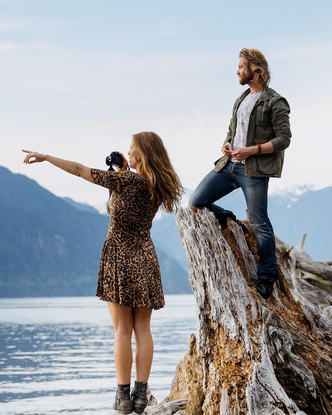 A couple poses on driftwood overlooking a lake and mountains. A woman is holding a camera and pointing to the left.