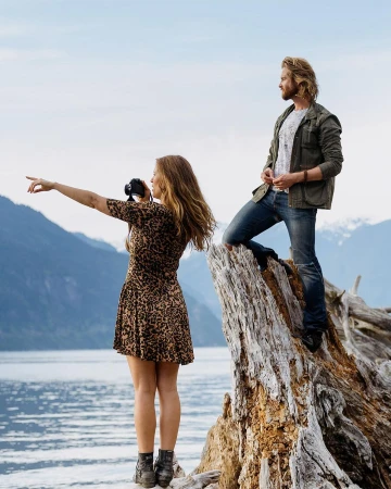 A couple poses on driftwood overlooking a lake and mountains. A woman is holding a camera and pointing to the left.