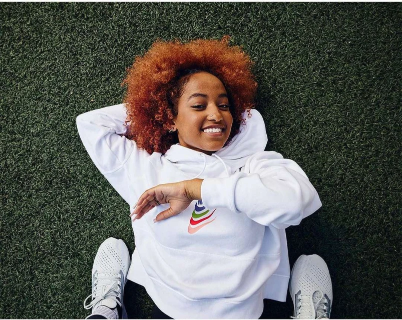 Smiling young woman with red hair in white Nike hoodie lies on green turf grass with arms above head, wearing Nike sneakers. Overhead shot.