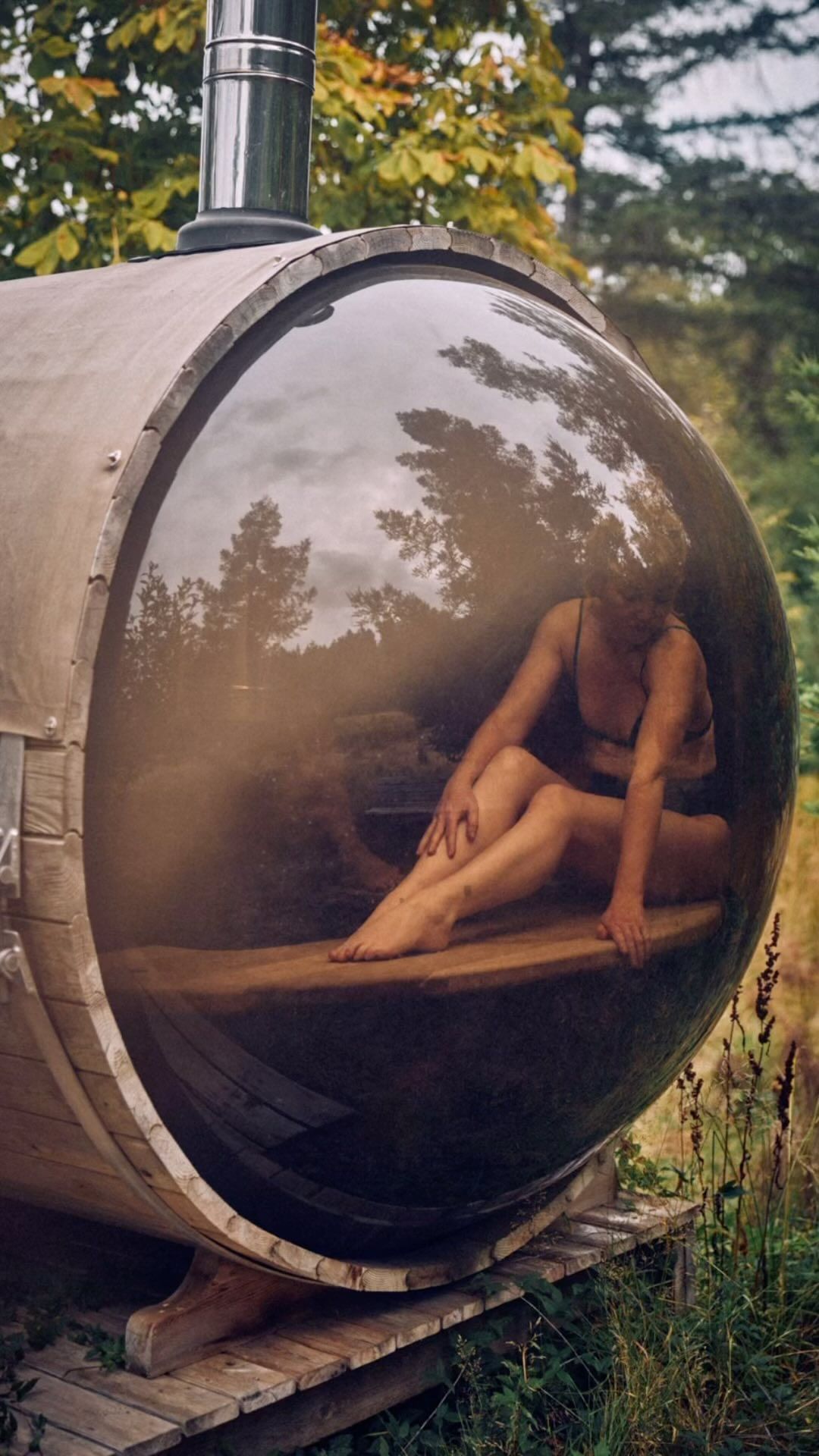 A woman sits inside a barrel-shaped sauna outdoors, with natural light highlighting the structure and the surrounding green trees.