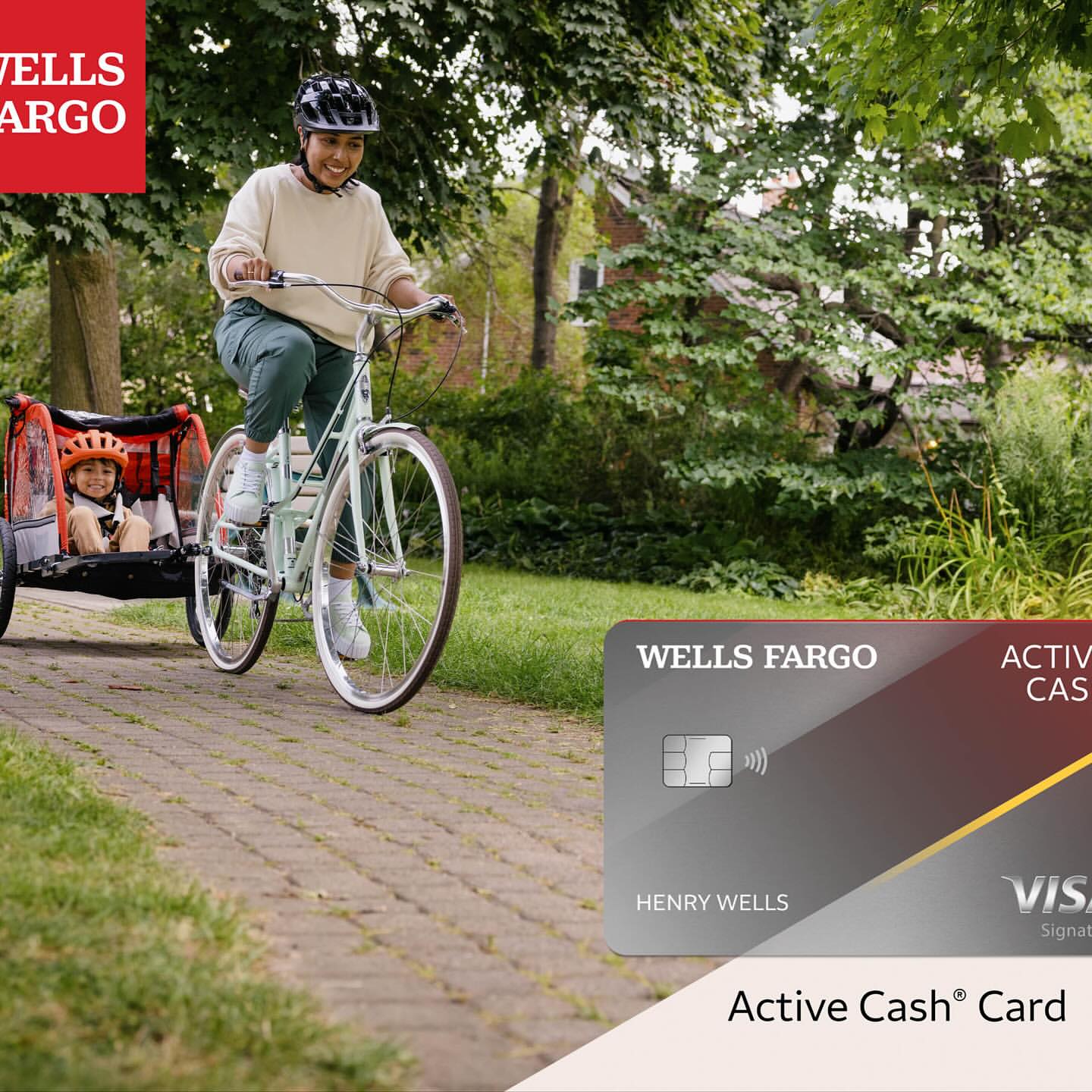 A woman in a helmet rides a bicycle, pulling a trailer with a child on a paved path lined with trees, next to a Wells Fargo credit card.
