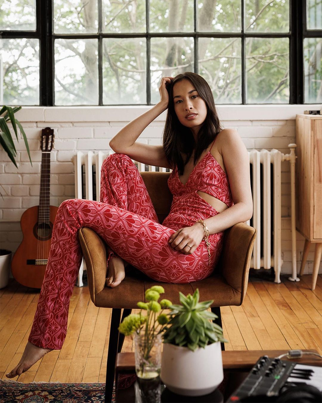 Adult woman sits in a chair wearing patterned red pants and top, with a guitar and plants visible indoors by a window.