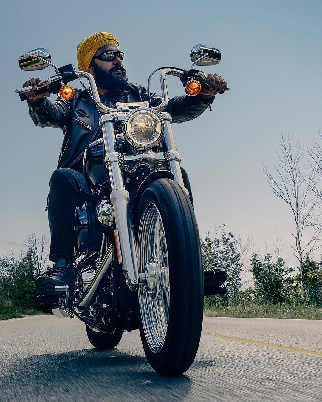 Man with a beard and yellow turban rides a black Harley Davidson motorcycle on a grey asphalt road with trees in the background.