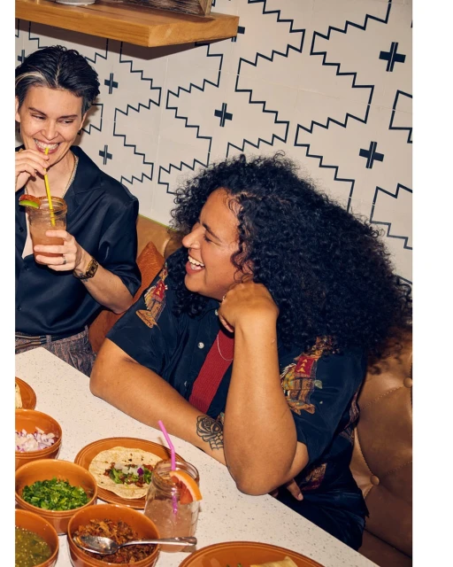 Two women are seated at a table in a restaurant with food and drinks, smiling and talking, in a brightly lit interior setting.