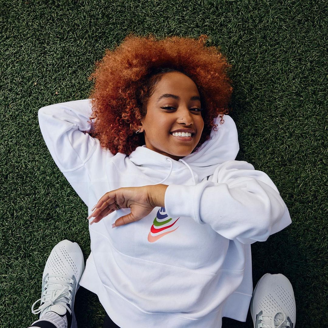 Smiling woman with auburn curly hair, wearing a white hoodie with a colorful logo, lying on her back on bright green grass.