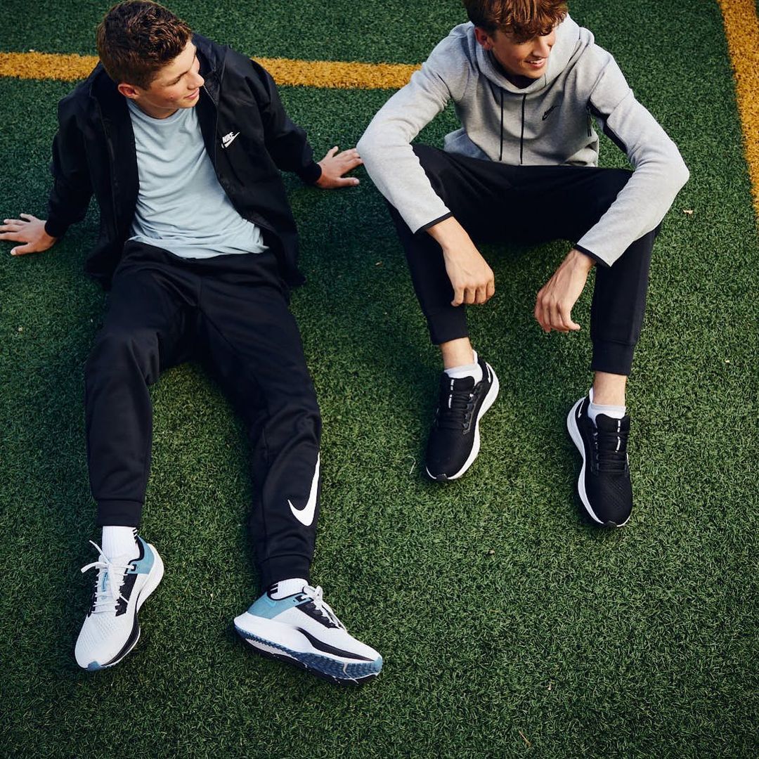 Two young men sit on a green field wearing athletic wear and sneakers. Both are looking away from the camera in a relaxed pose.