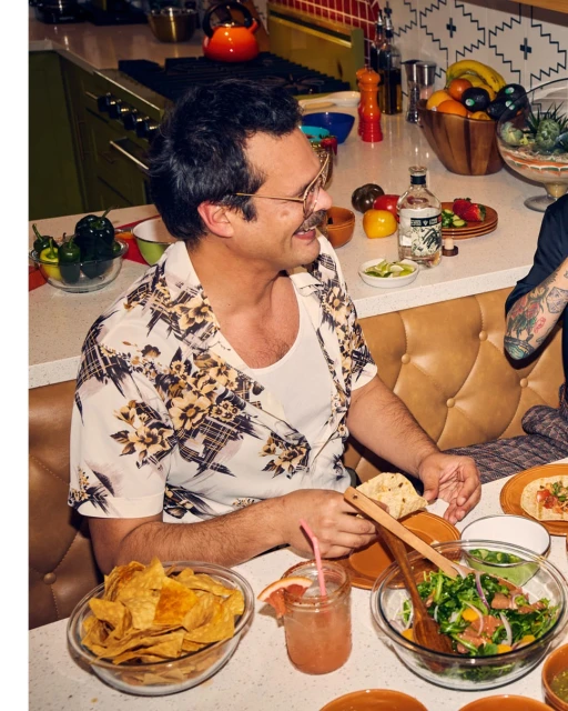 Man with mustache smiles at a table set with tortilla chips, salad, tacos, and a grapefruit drink at indoor dining setting.