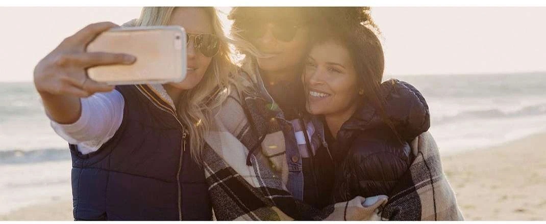 Three friends take a selfie together on the beach, wrapped in a blanket, with the ocean and sky visible in the background.