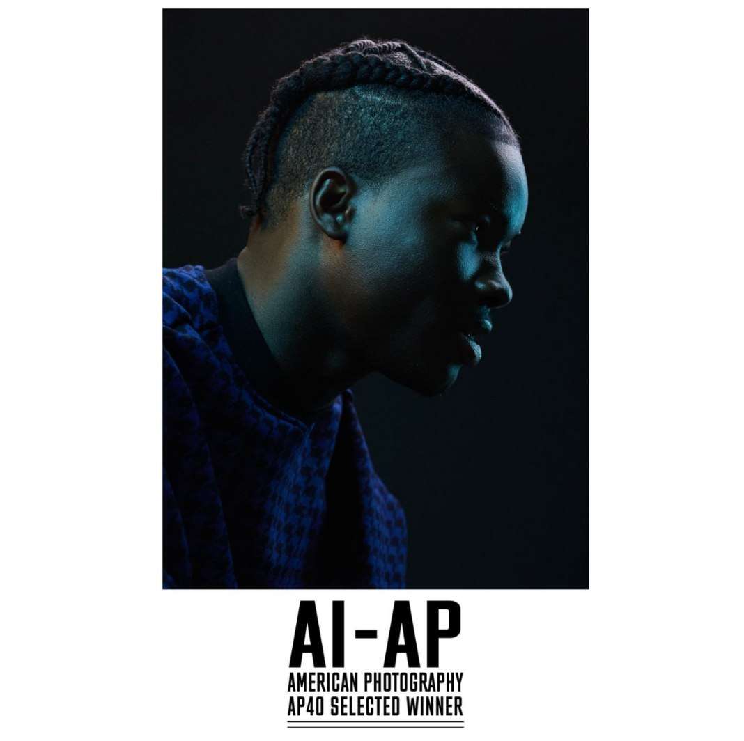 Portrait of a dark-skinned man in a blue patterned shirt with braided hair, set against a dark background with blue and white lighting.