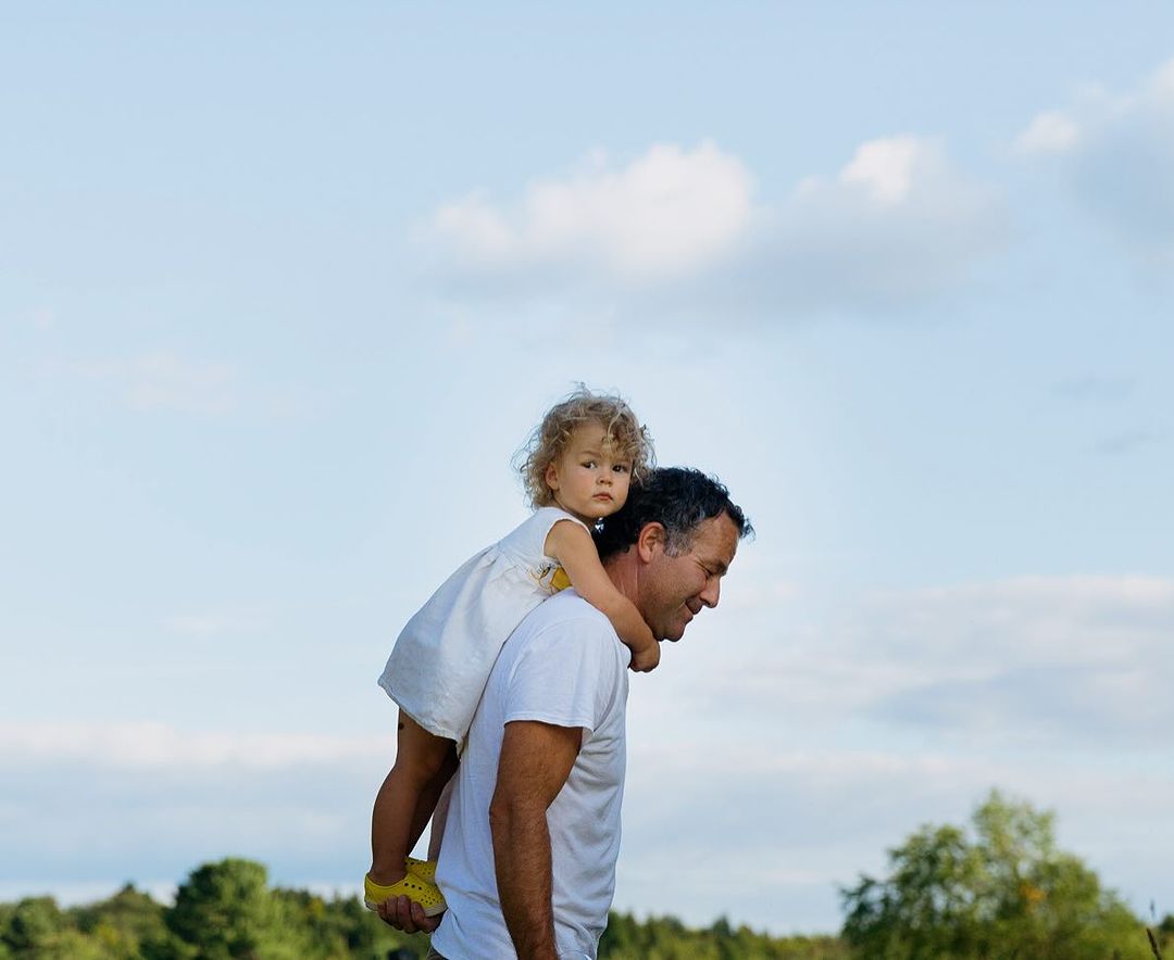 A father carries his young daughter on his shoulders in a daytime outdoor scene with a blue sky and green trees in the background.