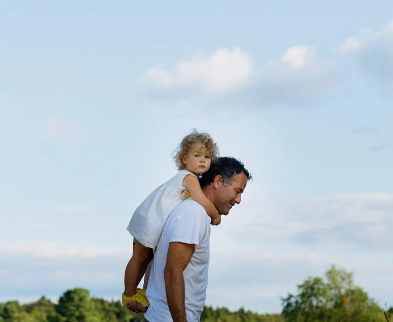 A father carries his young daughter on his shoulders in a daytime outdoor scene with a blue sky and green trees in the background.