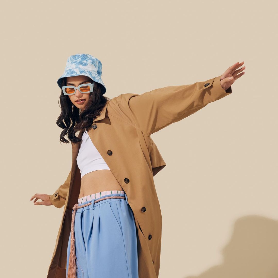 A stylish woman in a tan trench coat, bucket hat, and sunglasses poses in a studio with arms outstretched against a cream background.