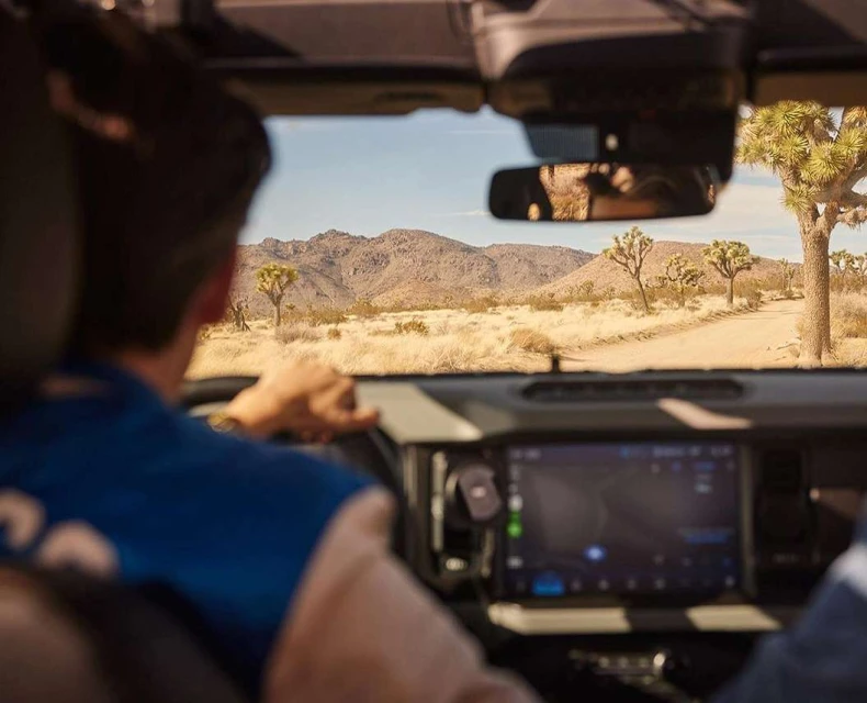 Man drives a car through the desert on a sunny day, the landscape features mountains and Joshua trees, and a digital dashboard is visible.